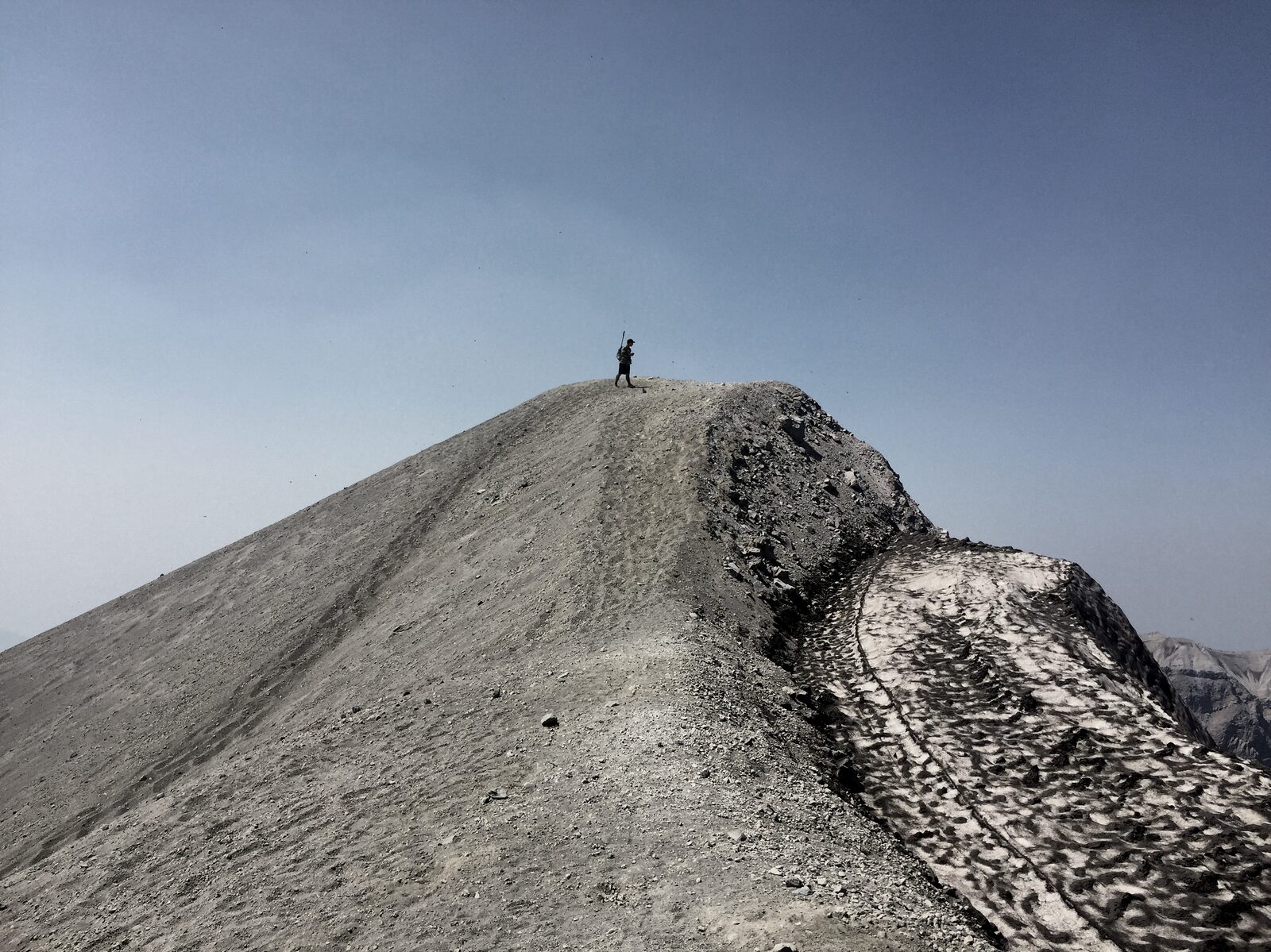 A single figure walking across a wide ash-pocked snowfield textured with sun-cups, an exposed ridge of dark volcanic rock to the right and distant peaks beyond