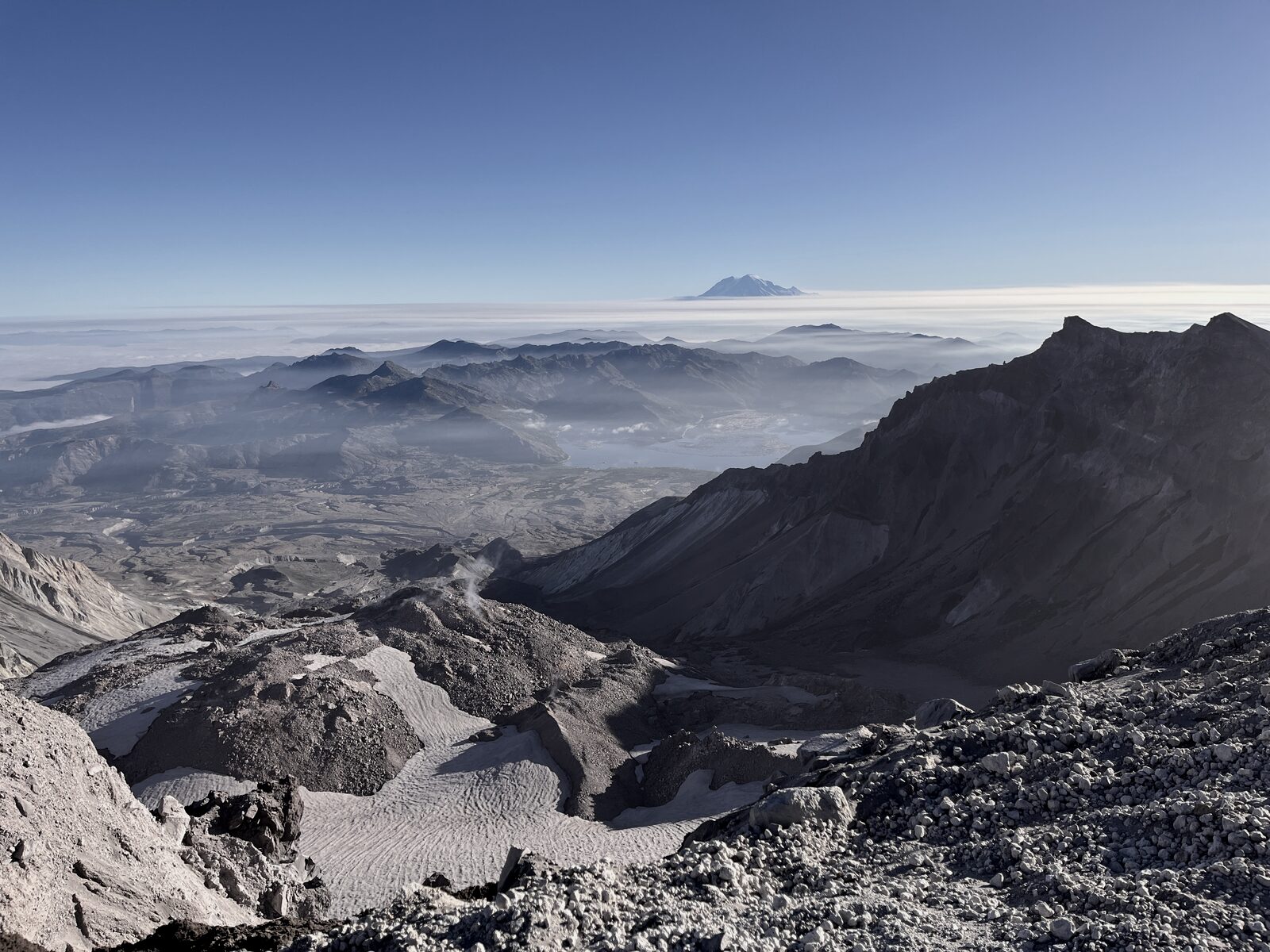 View from the rim of Mount St. Helens crater: ash and rock in foreground, marine cloud below, distant peaks on the horizon