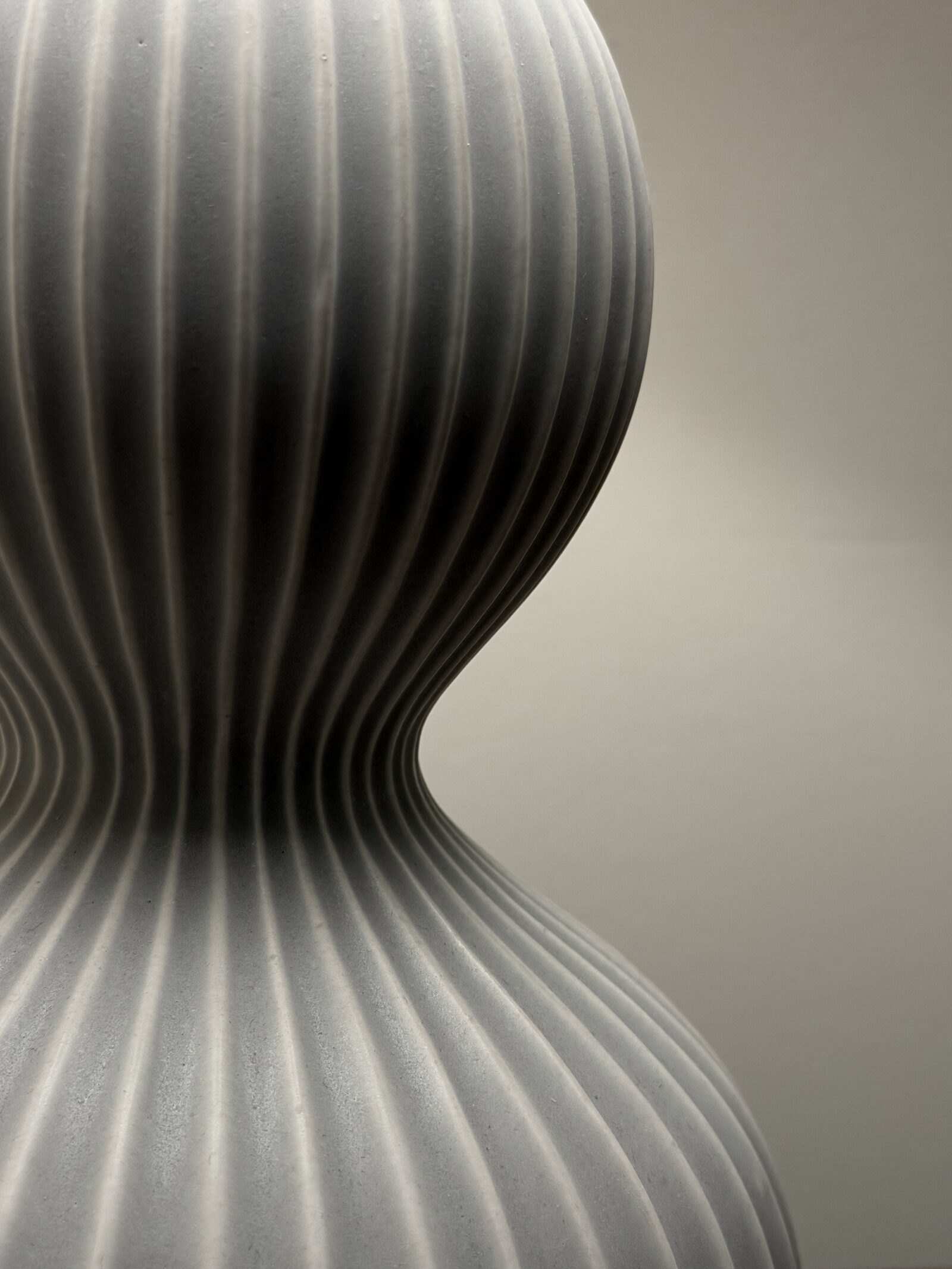 Close-up of a curved ribbed ceramic vessel with a pinched waist, in soft monochromatic gray light